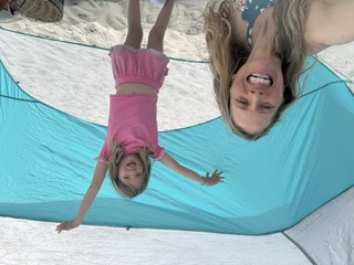A child with arms raised under a blue beach canopy beside a laughing guide
